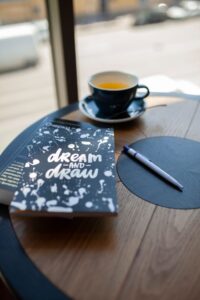 A notebook, pen and cup of tea on a wooden table by a window